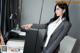 A woman in a business suit pointing at something on a desk.